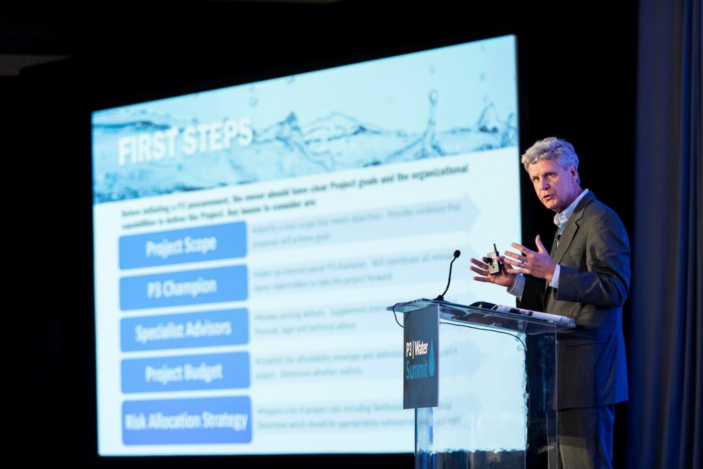 man speaking at lectern with large presentation screen behind him